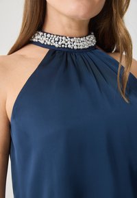 Sötétkék pánt nélküli felső sima anyagból; gyöngyökkel díszített gallérral és ráncolt részletekkel rendelkezik.