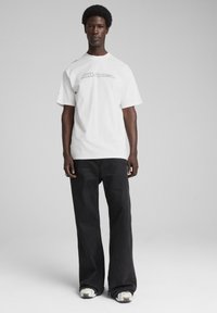 Hvid t-shirt med minimalistisk logo, korte ærmer, parret med sorte vide benbukser. Fodtøjet har en tekstureret sål med farvede detaljer.