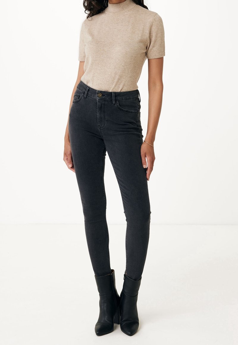 Black high-waisted skinny jeans with a button closure, featuring a smooth texture and fitted design. Paired with a beige short-sleeve top.
