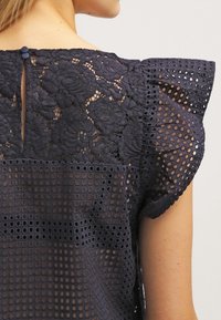 Blouse bleu marine à superposition de dentelle avec motif floral, épaules arrondies et dos à ouverture en forme de goutte avec fermeture par bouton. Texture en maille ouverte.