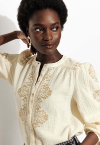 Blouse blanche à boutons avec broderie florale dorée, manches froncées, tissu texturé et col rond. Le modèle adopte une pose neutre.