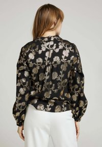 Blouse noire avec des broderies florales métalliques dorées, manches longues et un léger peplum à la taille. Texture lisse avec une coupe décontractée.