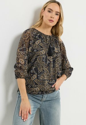 Kvinde iført sort og brun bluse med paisley-mønster og trekvartlange ærmer, kombineret med lyseblå jeans, stående foran en hvid væg.