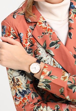 Femme portant un col roulé blanc et un blazer orange à motifs floraux, montrant son poignet avec une montre à cadran blanc et bracelet foncé.