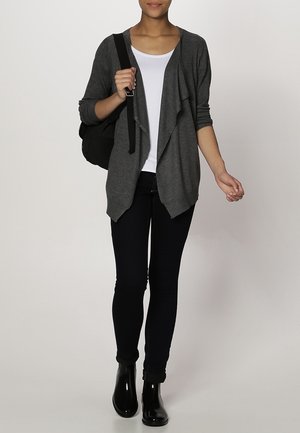 Femme portant un cardigan gris ouvert, une chemise blanche, un jean skinny noir, des bottines noires, portant un sac à dos noir sur une épaule.