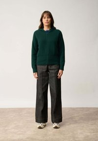 Zielona bluza z zamkiem, wykonana z miękkiego materiału, w połączeniu z luźnymi ciemnoniebieskimi dżinsami. Ubranie posiada kaptur i wyraźne szwy.