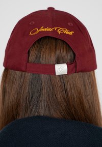 Bordeauxrote Kappe mit gelber stickierter "Special Club"-Aufschrift, ausgestattet mit einem Stoffriemen und einer metallischen Schnalle auf der Rückseite. Weicher Stoff.