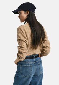 Beige Kabelstrickpullover mit umgeschlagenen Ärmeln, kombiniert mit hochtaillierten blauen Denim-Jeans. Das Modell trägt eine marineblaue Baseballkappe.