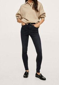 Unos pantalones vaqueros skinny de color azul oscuro combinados con un suéter de punto beige claro que tiene un cuello y tres botones, complementados con unos mocasines negros.