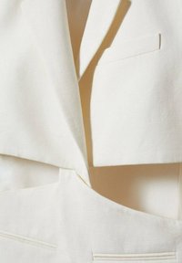 Gros plan sur une veste blazer blanc cassé avec un revers cranté, une poche poitrine et un détail assorti à la ceinture.