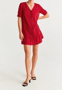 Czerwona sukienka krótki rękaw z dekoltem w serek, przednim zapięciem na guziki i falbaną u dołu. Wykonana z gładkiego materiału. Połączona z czarnymi sandałami na obcasie.