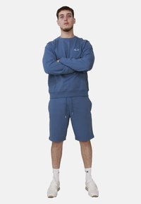 Felpa blu in cotone con collo a giro e logo, abbinata a pantaloni corti in cotone coordinati. Il modello è in piedi con le braccia incrociate, indossando sneakers bianche.