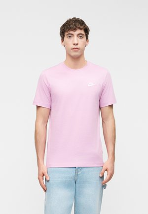 Joven con cabello rizado que lleva una camiseta rosa claro de Nike y pantalones vaqueros azul claro, de pie contra un fondo gris liso.