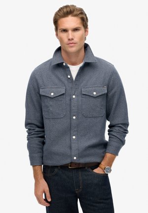 Mand iført en grå skjortejakke med klaplommer, hvid t-shirt, mørke jeans, brun bælte og armbåndsur med brun læderrem.