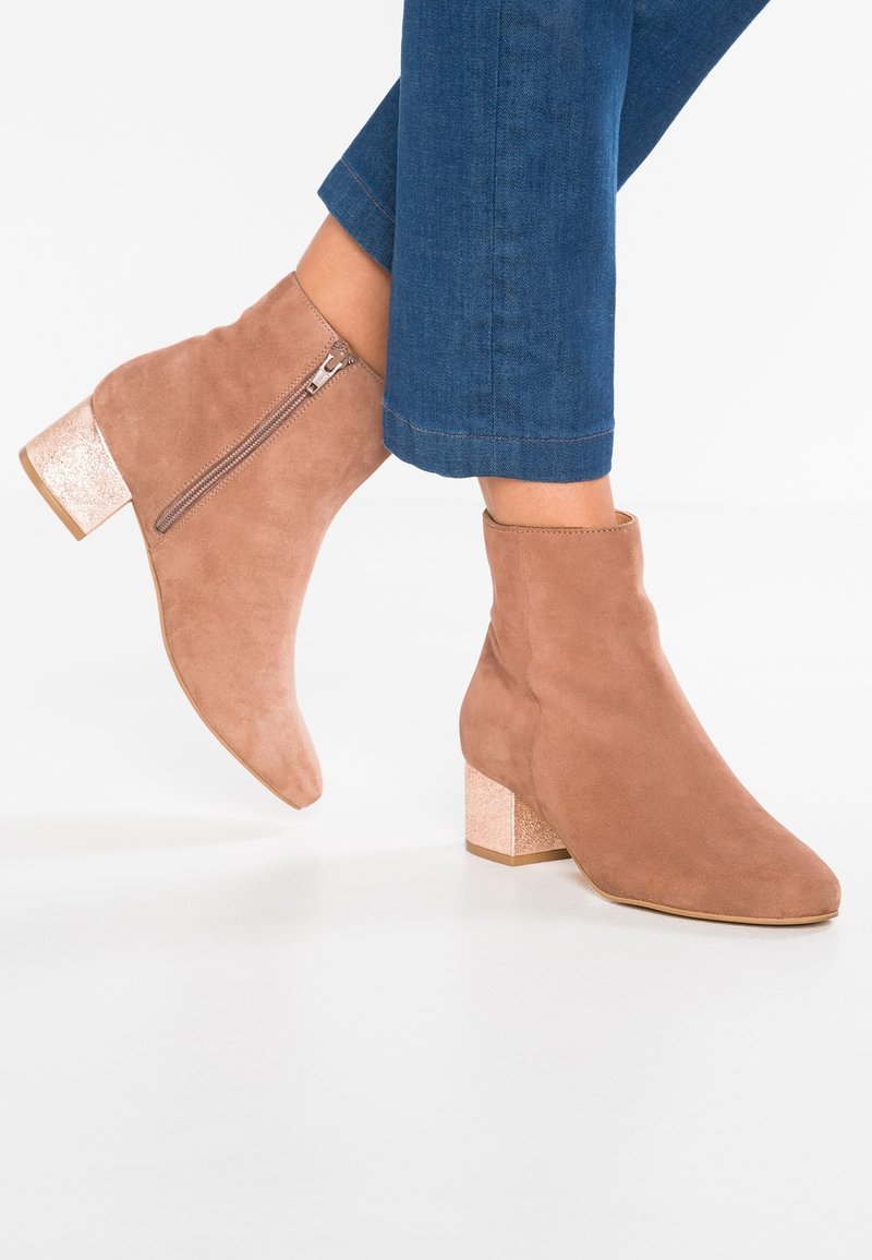 Bottes en suède marron clair montant à la cheville, avec fermeture éclair latérale et talons blocs métalliques, portées avec un jean bleu court.