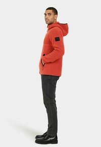Fluffig orange hoodie med svarta detaljer, framfickor med dragkedja, elastiska manschetter och huva. Bärs med mörkgrå byxor och svarta skor.