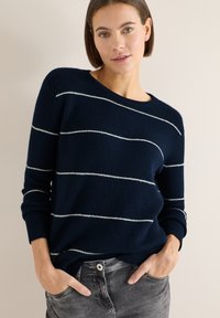 Navy striped sweater with white horizontal lines, ribbed texture, round neckline, and long sleeves, paired with grey jeans.