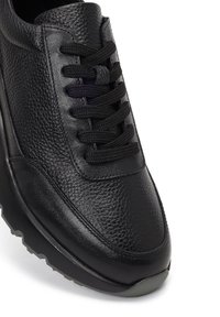 Zwarte leren sneaker met een gestructureerde bovenkant, ronde neus, zwarte veters en een dikke rubberen zool met een subtiel profiel.