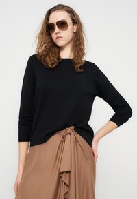 Μαύρο πλεκτό πουλόβερ με στρογγυλή λαιμόκοψη, μακριά μανίκια και λεπτομέρεια με κόμπο στο πλάι, συνδυασμένο με καφέ φούστα που έχει βολάν στο τελείωμα.