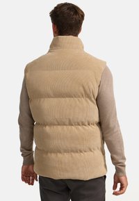Beige Puffer-Weste mit hohem Kragen, gerippter Textur und gepolsterten Abschnitten. Über einem hellbraunen Langarmshirt getragen.