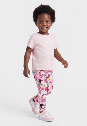 Copil zâmbitor purtând un tricou roz deschis, colanți colorați cu modele și adidași roz luminoși, mergând pe un fundal alb simplu.