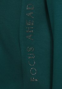 Türkiser Stoffoberteil mit der Aufschrift "FOCUS AHEAD", verziert mit silbernen Strasssteinen an der Seite, weiche Textur, lässiger Schnitt.