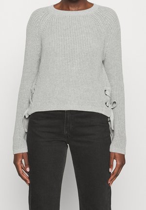 Pull en maille gris clair avec des liens sur les côtés, porté avec un jean noir, montrant le ventre et les bras sur un fond uni.