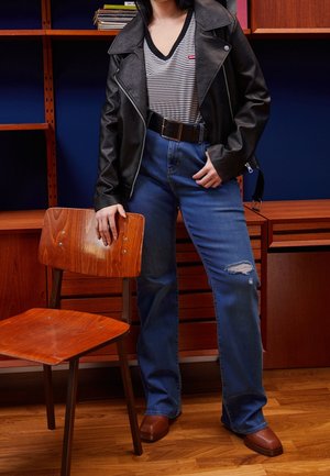 Person iført sort læderjakke, striped skjorte, revnede blå jeans og brune sko, stående ved siden af en træstol og et skrivebord i et rum.