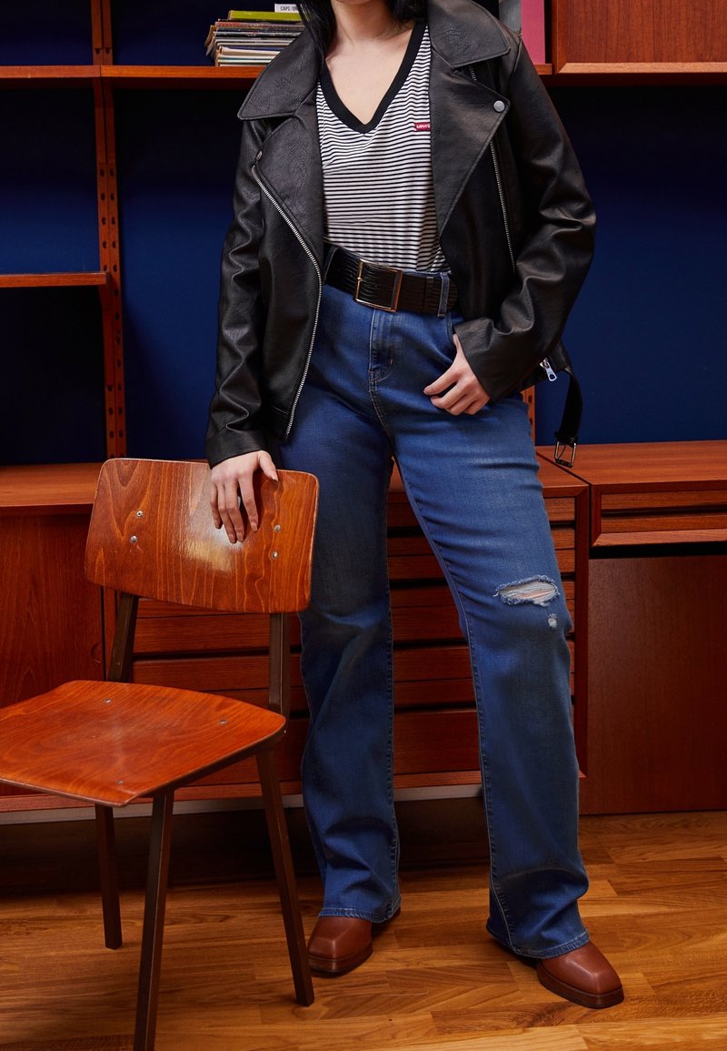 Personne portant une veste en cuir noire, une chemise rayée, un jean bleu déchiré et des chaussures marron, debout à côté d'une chaise en bois et d'un bureau dans une pièce.