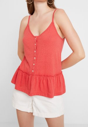 Vrouw in een rode mouwloze blouse met knopen en een rimpelrand, met witte shorts, staand tegen een effen lichte achtergrond.