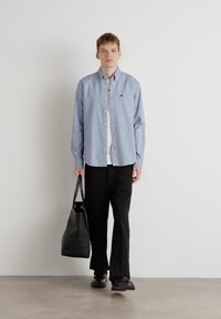 Lys blå skjorte med knapper, hvit t-skjorte, svarte bukser, bærende en svart totebag, iført svarte sko, stående mot en enkel vegg.