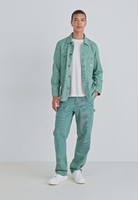 Veste vert menthe texturée avec des coutures et des boutons visibles, assortie à un pantalon correspondant avec poches et coupe décontractée. T-shirt blanc en dessous.