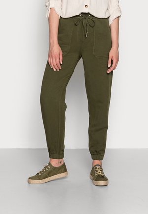 Modell trägt olivgrüne Hosen mit Kordelzug, vorderen Taschen und passend grüne Schnür-Sneaker auf grauem Boden.