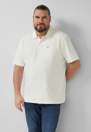 Mann mit Bart und zurückgebundenen Haaren, trägt ein weißes kurzärmeliges Poloshirt und blaue Jeans, steht vor einem grauen Hintergrund.