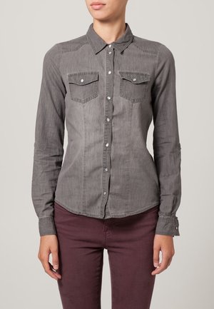 Chemise grise à boutons avec deux poches poitrine, manches longues et ourlet arrondi. Le tissu semble doux avec une texture subtile.