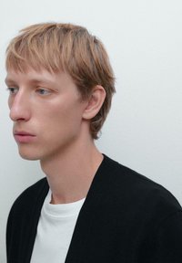 Svart cardigan över en vit t-shirt, med en avslappnad passform och minimalistisk design. Håret är ljusbrunt, stylat rakt med en lätt texturerad look.