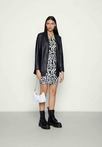 Blazer de pele preta sobre um vestido com padrão de espirais brancas e pretas. Botas pretas com atacadores e uma pequena mala branca com uma alça de corrente.