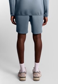 Shorts di cotone azzurro chiaro con una texture liscia, abbinati a sneaker beige e calzini bianchi. Cuciture visibili e un design a taglio rilassato.