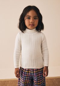 Pull en maille blanche à col montant, avec un motif torsadé ; associé à un pantalon à carreaux colorés rose, jaune et bleu.