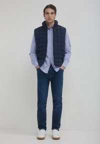 Gilet trapuntato blu navy sopra una camicia azzurra con colletto, abbinato a jeans blu scuro e sneakers bianche. Texture lisce, linee pulite e design semplice.