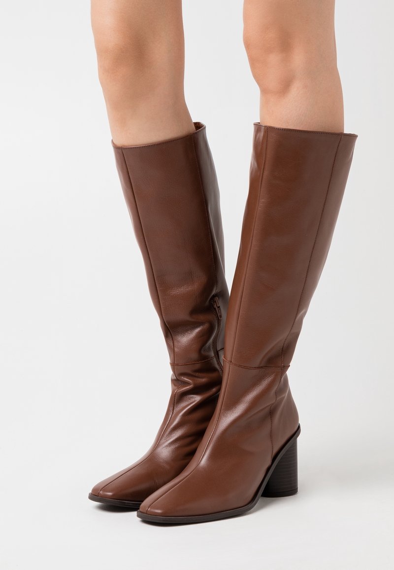 Botas de cuero marrón hasta la rodilla con un diseño desestructurado, punta cuadrada y un tacón grueso. Textura suave con detalles de costura sutiles.