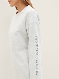 Külgvaade inimesest, kes kannab heledat halli pika varrukaga pluusi, millel on varrukal vertikaalselt trükitud "Tom Tailor".