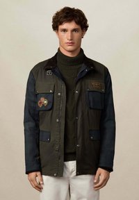 Mladić u tamnozelenoj i mornarskoj jakni s aplikacijama, zelenoj rolci i bež hlačama, stoji nasuprot jednostavne pozadine.