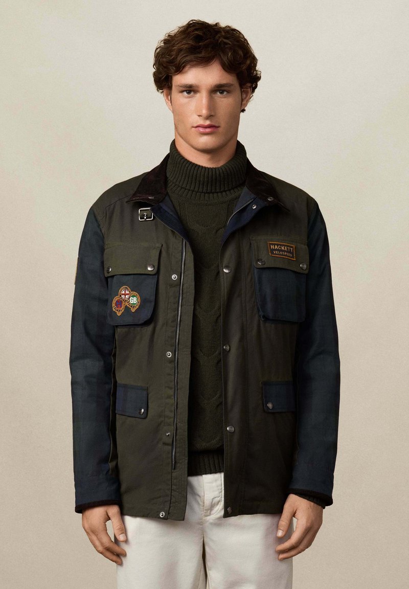 Mladić u tamnozelenoj i mornarskoj jakni s aplikacijama, zelenoj rolci i bež hlačama, stoji nasuprot jednostavne pozadine.