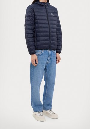 Homme portant une veste matelassée noire EA7 Emporio Armani, un jean bleu clair et des baskets écrues, debout devant un fond uni clair.
