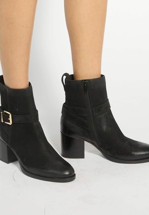 Bottines en cuir noir avec talons carrés, fermeture éclair latérale et bride décorative avec boucle dorée, portées aux pieds d’une personne.
