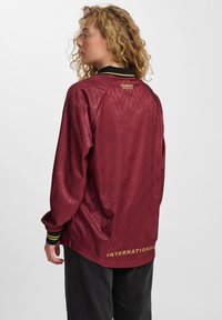 Kihara naise, kes vaatab külgvaates, kannab tumepunast pika varrukaga spordiriiete toppi, mille tagaküljel on "INTERNATIONAL" tekst.