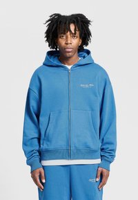 Modrá mikina s kapucí na zip vyrobená z měkké tkaniny, s přední kapsou a žebrovanými manžetami. Na hrudi má výšivku loga.