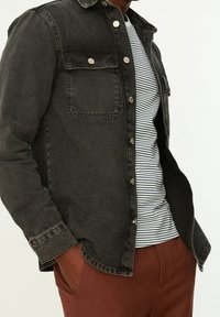 Camicia di denim nero con due tasche sul petto con bottoni, dettagli in argento e orlo curvo, indossata sopra una t-shirt a righe bianche e blu navy.