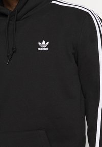 Schwarzer Hoodie mit weißem Adidas-Logo und seitlichen Streifen. Verfügt über eine Kapuze mit Kordelzug und eine Kängurutasche. Hergestellt aus weichem Stoff.